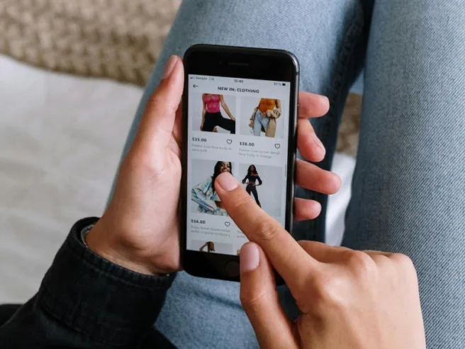Shopping på mobilen. En hånd holder en telefon mens en annen hånd trykker på skjermen, som viser en nettbutikk som selger klær.