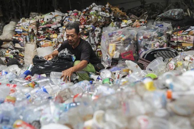 En mann sitter blant et hav av plastflasker og annet søppel